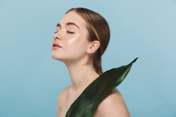 Beautiful young woman take care of her skin isolated over blue wall background posing with leaf...