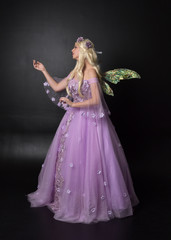  full length portrait of a blonde girl wearing a fantasy fairy inspired costume,  long purple ball gown with fairy wings,   standing pose  on a dark studio background.