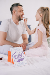 cute child applying lipstick on fathers lips near gift box and fathers day greeting card on bedding