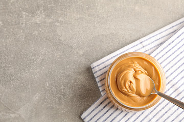 Glass jar with peanut butter and spoon, and kitchen towel on grey background, space for text and top view