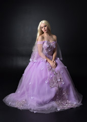 full length portrait of a blonde girl wearing a fantasy fairy inspired costume,  long purple ball gown with fairy wings,   sitting pose  on a dark studio background.
