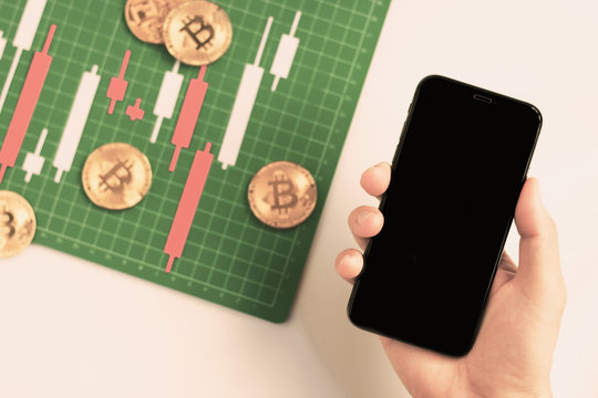 Top view hand holding smartphone with Candlestick chart make from color paper white and red on Green board with grid lines, investment, trading, stock, finance concept