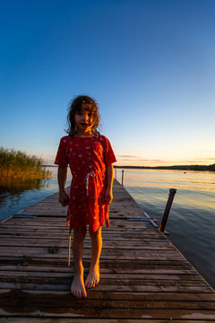Backlit summer night portrait of a young caucasian girl in red dress on a jetty with water and blue sky.