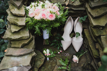 Botanic bridal chic. Bouquet with silk ribbons, female classic shoes, boutonniere, wedding rings and engagement ring on natural stone background.