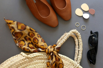 Fashionable accessories: sunglasses, straw bag with leopard print scarf, vintage brown leather shoes, gold rings and earrings on gray background. Flat lay.