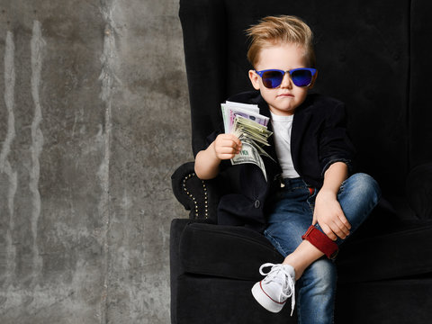 Portrait of self-confident rich kid boy siting in luxury armchair with bundles of dollars cash in his hand at text space