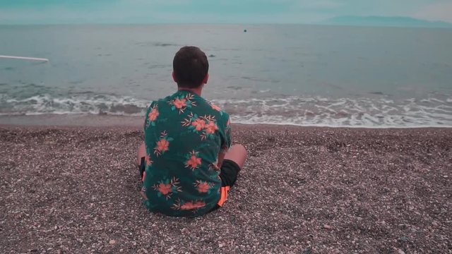 Young man sitting on the beach and  throwing pebbles into the sea. Alone man wearing floral shirt. Sunset in summer