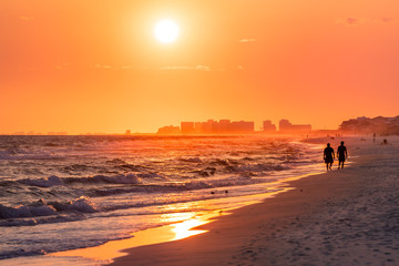 Fototapeta na wymiar Couple walking on shore with dramatic orange red yellow sunset in Santa Rosa Beach, Florida with Pensacola coastline coast cityscape skyline in panhandle with ocean gulf mexico waves
