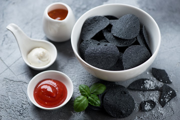 Fototapeta na wymiar White bowl with black potato chips and dipping sauces over grey concrete background, studio shot