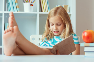 Portrait of funny little school girl relaxing reads book at the table in room at home