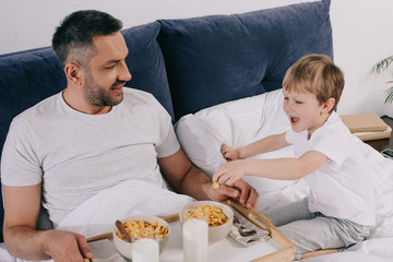 cheerful father and son having breakfast in while staying in bed together