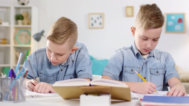 Chest-up shot of blond identical twin brothers, dressed in denim shirts, sitting side by side at table at home and doing homework, one boy writing in his exercise book, and another drawing