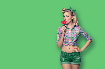 Photo of young woman eating heart shape lollipop, dressed in pinup style, over green color...