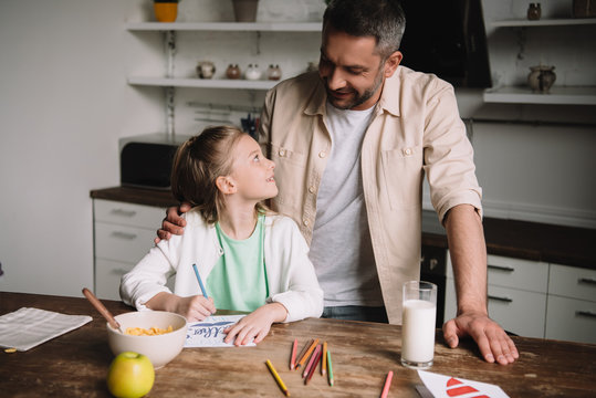 happy father hugging adorable daughter sitting at wooden table with served breakfast and drawing fathers day greeting card