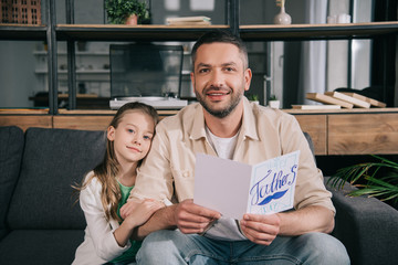 cute daughter smiling father holding fathers day greeting card and smiling at camera