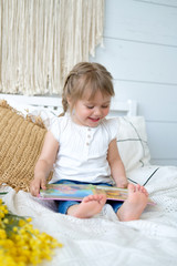 Beautiful little girl sitting on the bed and reading a book. The baby has blonde hair and blue eyes. White bedroom, cozy house. Teaching children to read.