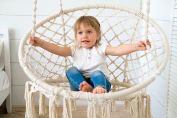 Beautiful little girl swinging in a hanging chair. The baby has blonde hair and blue eyes. White bedroom, cozy house. The interior design of the nursery concept