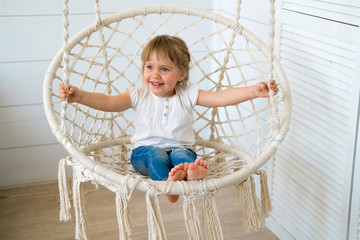 Beautiful little girl swinging in a hanging chair. The baby has blonde hair and blue eyes. White bedroom, cozy house. The interior design of the nursery concept