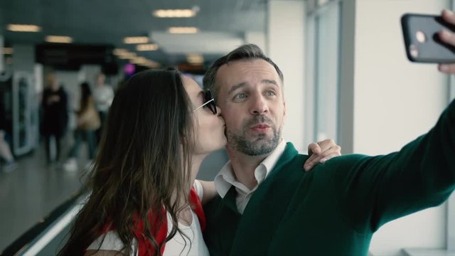 Young beautiful couple in the hall of airport. Brunette smiling woman kissing man in cheek, while taking selfie photo on smartphone near the window. Cinematic shot on RED camera.