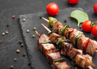 Grilled pork and chicken kebab with paprika on stone chopping board with salt, pepper and tomatoes on black background.