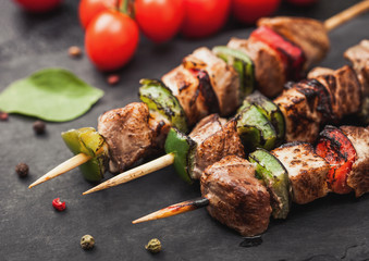 Grilled pork and chicken kebab with paprika on stone chopping board with salt, pepper and tomatoes on black background.