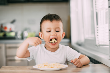 a child in the kitchen during the day eating pasta in a spiral in a white t-shirt