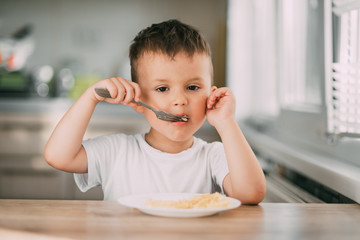 a child in the kitchen during the day eating pasta in a spiral in a white t-shirt