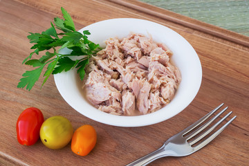 Canned tuna fillet in white porcelain bowl on a brown wooden cutting board with fork, parsley and three cherry tomatoes. Seafood, healthy eating.