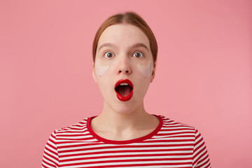 Portrait of cute young amazed red-haired lady in a red striped T-shirt, with red lips and with patches under the eyes, looks at the camera with wide open mouth and eyes stands over pink background.