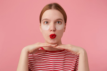 Close up of funny cute young happy red-haired lady in a red striped T-shirt, with red lips, leaning the chin on the hands and very pleased with new patches. Stands over pink background.
