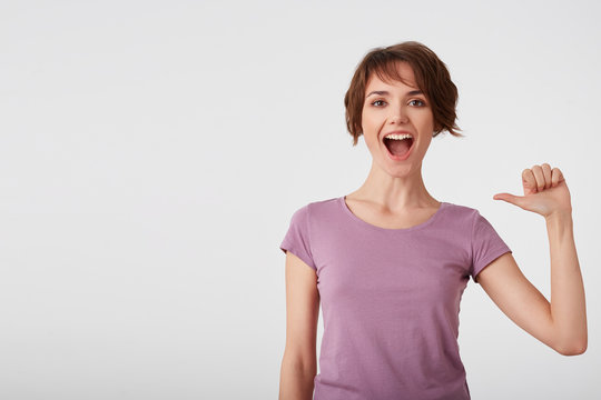 Self assured cheerful short-haired lady in blank t-shirt feels proud of her deeds, points at herself feels surge of pride, raises head, broadly smiles, wears casual clothes, isolated over white wall.