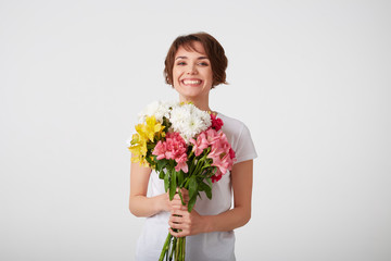 Portrait of smiling nice short haired lady in white blank t-shirt, holding a bouquet of colorful flowers, very glad and smiling over white wall.