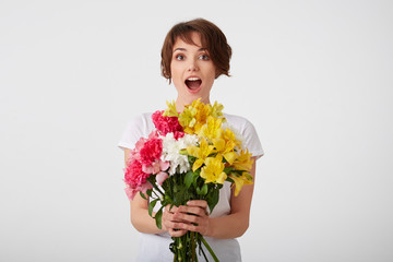 Wow, unbelievable, this flowers looks great! Happy young cute short haired girl in white blank t-shirt, with wide open mouth and eyes, holding a bouquet of colorful flowers, isolated over white wall.