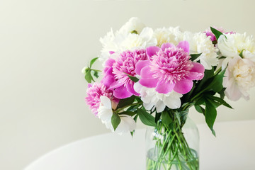 Close-up bouquet of fresh big pink, white and cream peonies in simple glass jar on glance table indoor. Vase with beautiful tender spring flowers on glass table