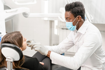 Obraz premium Doctor treats teeth. Young African male dentist with a patient. Woman in the dentist chair at dental clinic. Medicine, health, stomatology concept. dentist treating a patient.