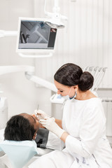 Young woman dentist treating root canals in the dental clinic. Young African American male with bad teeth lying on dentist chair with open mouth.