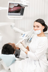 Inspection with a dental macro camera and displays the result on the monitor. Young woman dentist treating root canals in the dental clinic. Man patient lying on dentist chair with open mouth.