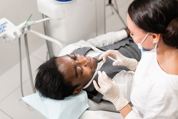 Obraz premium Young woman dentist treating root canals in the dental clinic. Young African American male with bad teeth lying on dentist chair with open mouth.