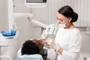 Young woman dentist treating root canals in the dental clinic. Young African American male with bad teeth lying on dentist chair with open mouth.