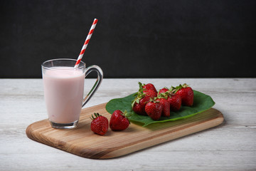 Homemade yogurt with fresh strawberry in transparent cup with colorfull stick on a wooden board. Fresh strawberries on a big green leaf near.