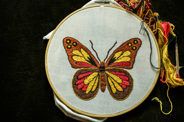 Bright butterfly embroidered on white cotton fabric stretched on a wooden hoop with scissors and...