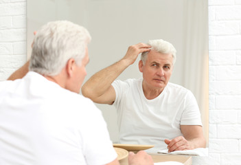 Senior man with hair loss problem looking in mirror indoors