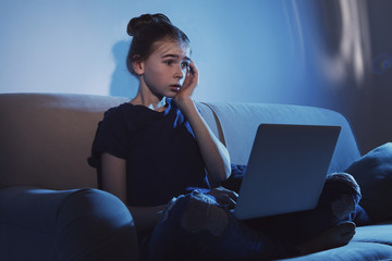 Shocked teenage girl with laptop on sofa in dark room. Danger of internet