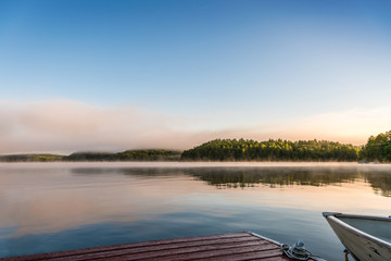 A early summer  morning mist lifting off a tranquil lake, to expose the forest across the lake with painterly reflection on lake, foreground dock and boat. copy space