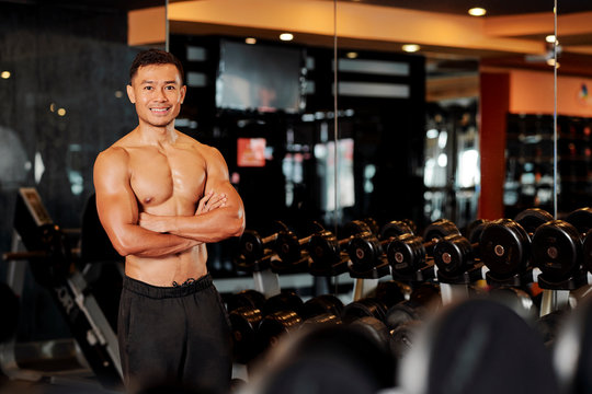 Portrait of confident shirtless fitness instructor standing in gym with his arms folded and smiling at camera