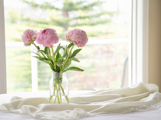 Transparent vase of pink peonies flower bouquet on white table in front of a window © Una