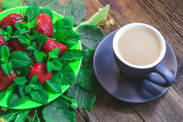 a cup of coffee with milk and strawberries, melissa leaves and mint in a plate on a wooden kitchen table.