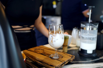 Iced latte - A glass of coffee with milk, topped with milk foam on wooden table and flower vase background, Refreshing Summer Drink.