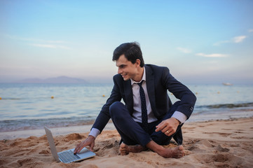 close up photo of a young man in suit with laptop working on the beach and talking to someone