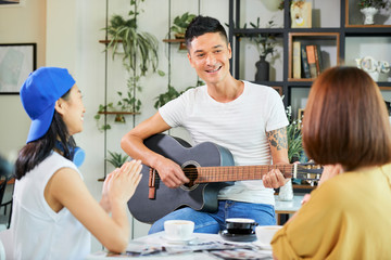 Positive young mixed-race man playing guitar and signing for his clapping friends